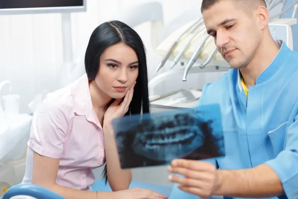 A woman holding her hand to her cheek while at the dentist while he shows her an xray of her teeth