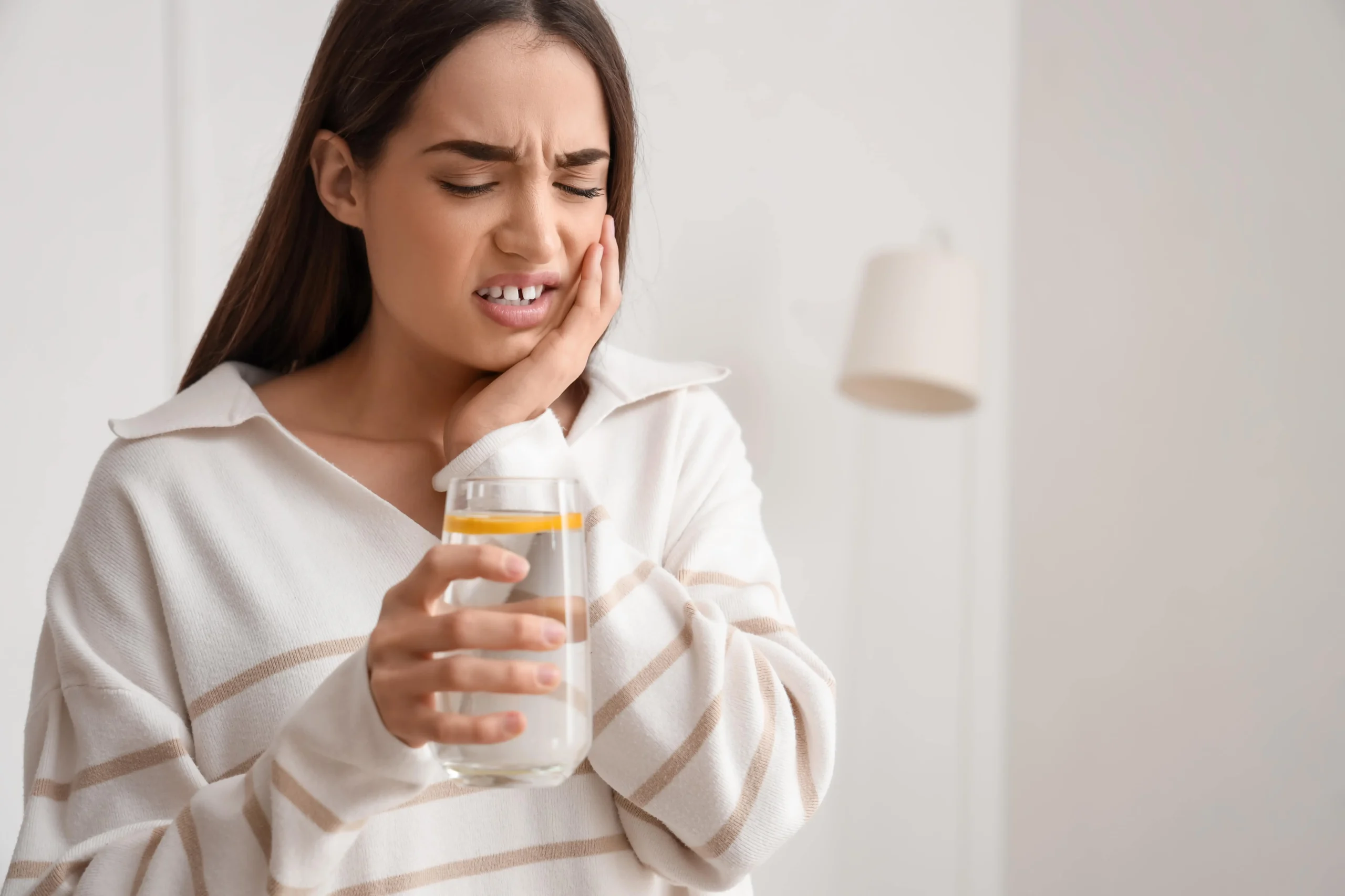 A woman holding her hand to her cheek while wincing in pain and holding a glass of water